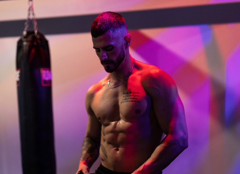 The coach stands in dramatic lighting in a gym near a heavybag.